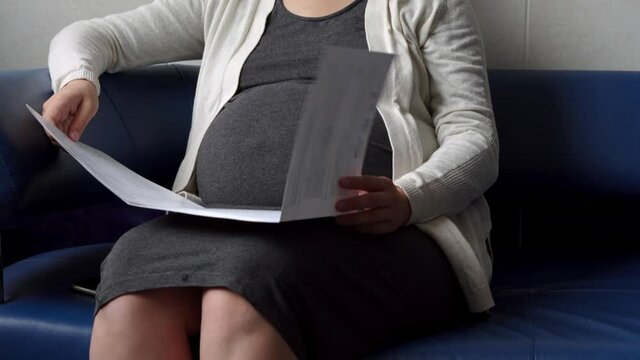 Young Pregnant Woman In Medicine Protective Mask Reading On Couch Waiting For Appointment Medical Control On Hospital. Visit Gynecologist Doctor At Clinic. Examine Expectant Belly Healthcare Check Up