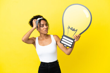 Young african american woman isolated on yellow background holding a bulb icon and having doubts