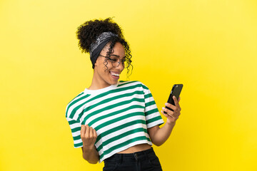 Young african american woman isolated on yellow background using mobile phone and doing victory...