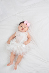 Smiling baby girl toddler in white dress and hair bow decoration lying on bed