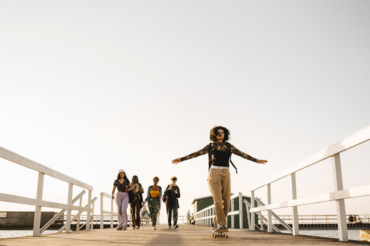 Teenage girl skateboarding while multiracial friends walking on pier against clear sky - Powered by Adobe