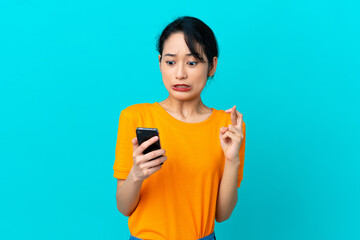 Young Vietnamese woman isolated on blue background using mobile phone with fingers crossing