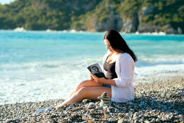 A girl sits on the seashore and reads a book.
