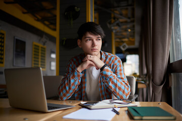 Pensive male student sitting front open laptop computer while looking away. Young man freelancer working remote  with online project on netbook