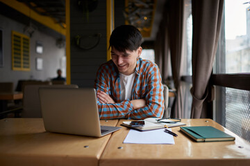 Positive male student sitting front open laptop computer while reading literature. Young man freelancer working remote  with online project on netbook