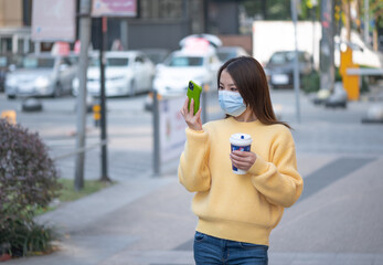 Beautiful Chinese Asian young woman walks on street during sunny autumn day. Holds drinks cup of coffee, takes pictures selfie using mobile cell phone. Wearing face mask to prevent respiratory disease
