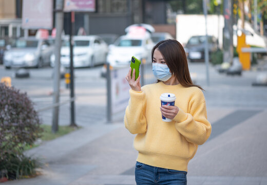 Beautiful Chinese Asian young woman walks on street during sunny autumn day. Holds drinks cup of coffee, takes pictures selfie using mobile cell phone. Wearing face mask to prevent respiratory disease