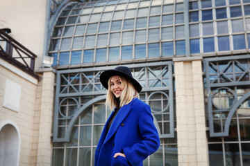 Beautiful young woman in black felt hat and bright blue coat looking at camera and smiling, on background beautiful building. Lifestyle portrait of happy female