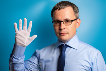 A serious man in a blue shirt and tie shows five fingers with his hand in a white glove. Confident specialist on a blue background.