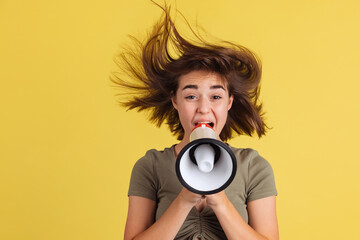 Half-length portrait of young girl, student shouting at megaphone isolated on yellow studio backgroud.