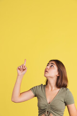 Vertical composition of portrait of young pretty girl in brown tee pointing up isolated on yellow studio backgroud.