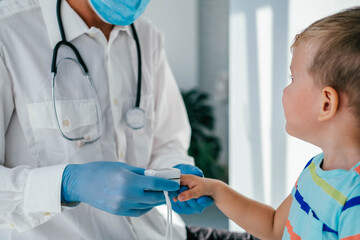 A doctor wearing medical gloves and a mask puts a pulse oximeter on the child's finger to measure...