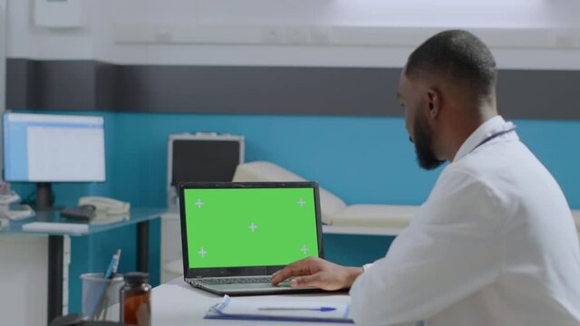 African american physician doctor looking at mock up green screen chroma key laptop computer with isolated display typing medical expertise working in hospital office. Medicine concept