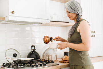 Mature asian woman with grey hair making coffee in kitchen