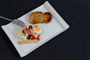 White plate of Russian salad made with mayonnaise, pepper, egg accompanied by bread on a black background, typical food in Spain