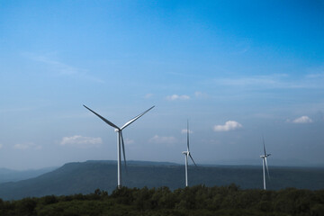 The big white wind turbine for spinning electricity sees large mountains far away, with clouds and blue skies above the mountains.