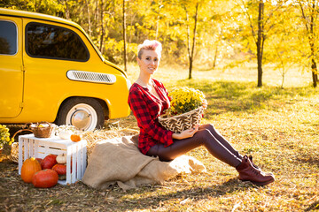 happy smiling beautiful woman with flowers and pumpkins in autumn park background near yellow car