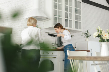 The older mother prepares food in the kitchen, the daughter drinks tea and looks at her mother