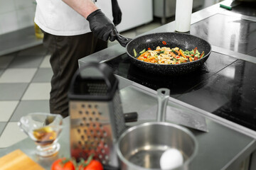 Close-up of the hands of a chef who is frying meat and vegetables in a pan in a restaurant
