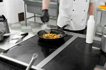 Close-up of the hands of a cook who is frying meat and vegetables in a pan and adding sauce or broth