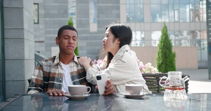 Young woman consoling her friend feeling frustrated after reading the worst news on a cell phone while sitting together in the rooftop on an outdoor cafeteria. Focus on emotions of desolation, despair