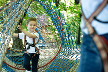 Portrait of a little boy in climbing gear in a rope park, holding a rope with a carbine.