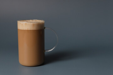 Glass cup of hot caramel cappuccino with bubbles on grey background.