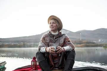 Portrait of woman dressed in poncho, sit in the boat, drink tea and look to the lake, enjoy life, happy in moment