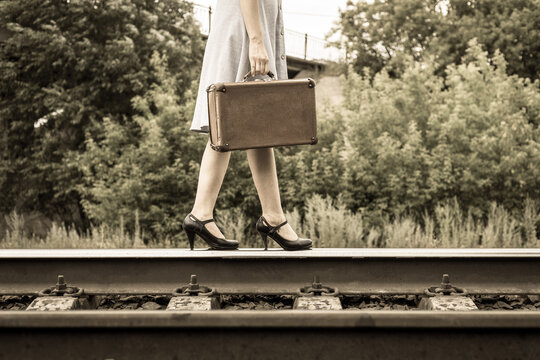 A young woman in a dress and black shoes walks along the train rails with an antique suitcase. Women's feet in high-heeled shoes walking on train rails, close-up.