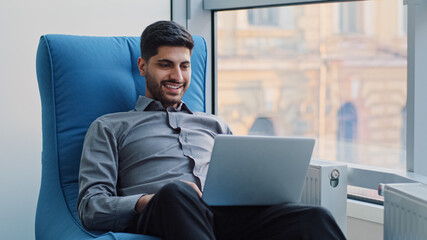 Happy millennial Indian corporate employee in creative profession sits in comfortable armchair in coworking space using computer. Young Arabic professional working on laptop, dreaming, create new idea