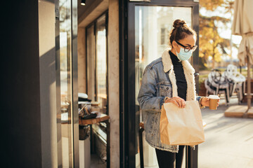 Young woman wearing protective face mask holding takeaway food and coffee to go, standing near restaurant window. Social distancing during quarantine caused pandemic.
