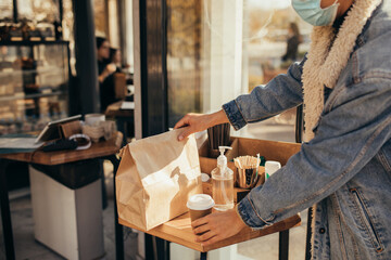 Young woman wearing protective face mask holding takeaway food and coffee to go, standing near...