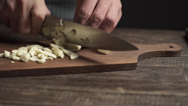 The chef in apron cuts the garlic with a knife in a rustic style on a dark background. Close-up