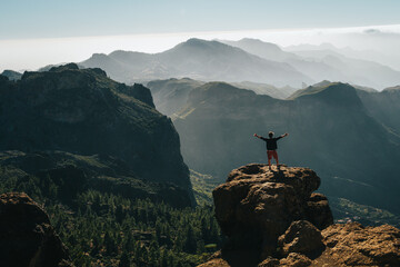 Man with arms outstretched on a cliff looking at the valley with mountains and forests in the background