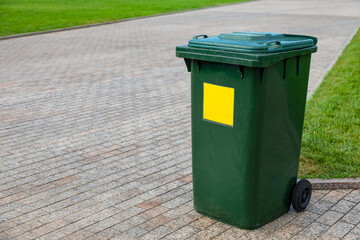 Plastic green garbage can for collecting garbage in the park. Mobile trash can with wheels. A yellow sign marks plastic waste