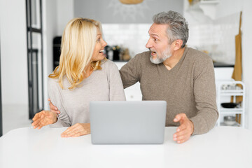 Shocked middle-aged couple feel euphoric sitting at the desk in front of laptop in the kitchen, looking at each other opened mouth, found out unexpected good news, got online sale offer or discount