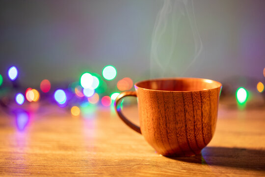 Wooden mug with coffee. Steam comes from the cup from hot coffee, chocolate, tea. Christmas lights in the background. Christmas is coming soon