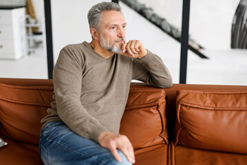 Portrait of thoughtful mid adult man sitting on the leather sofa at home. Handsome grey-haired man in casual wear relaxing on couch. Handsome mature bearded man looking away