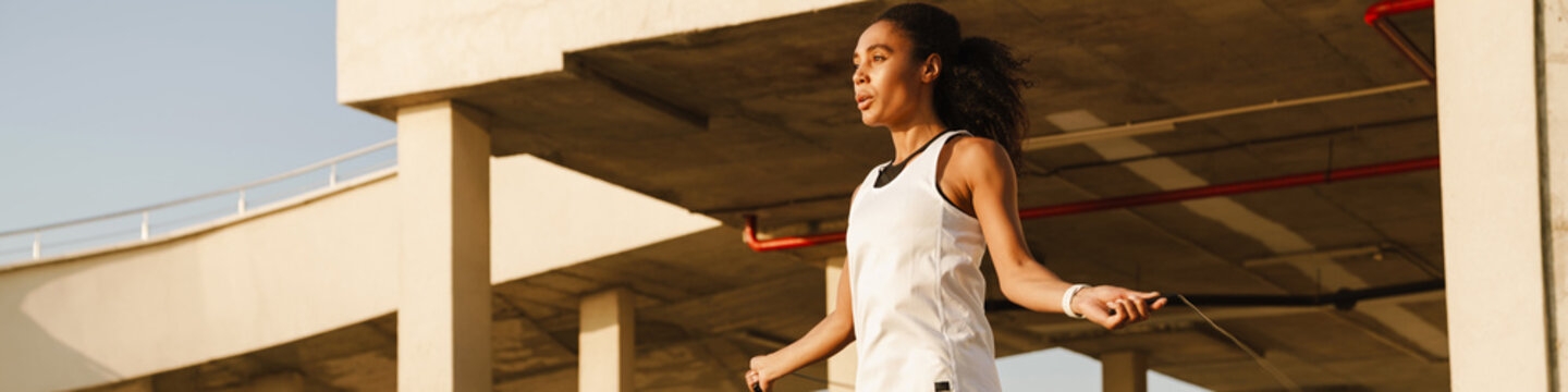 Black sportswoman working out with jumping rope on parking