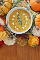 Bowl of butternut squash soup garnished with cream, rosemary, and chives, surrounded by various pumpkins, gourds, ornamental corn, and herbs on wood background with copy space.