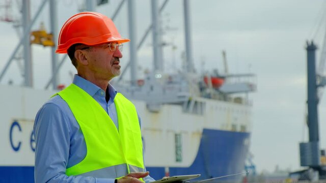 Sea cargo port industry. Middle aged foreman in uniform with tablet gives tasks to managee near large repairing vessel in dock closeup