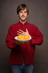 Young handsome tall slim white man with brown hair proudly presenting bowl of oranges in red shirt on grey background