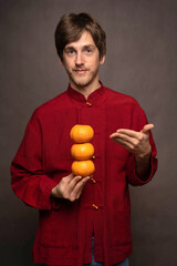 Young handsome tall slim white man with brown hair presentingly holding oranges in red shirt on grey background