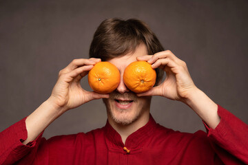 Young handsome tall slim white man with brown hair covering eyes with oranges in red shirt on grey background