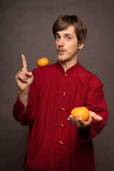 Young handsome tall slim white man with brown hair with tangerine on shoulder offering orange in red shirt on grey background