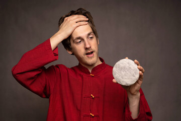 Young handsome tall slim white man with brown hair relieved holding pu erh tea cake in red shirt on grey background