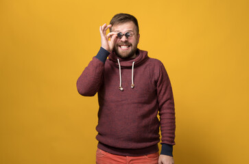 Cheerful Caucasian bearded man holds toy sunglasses like pince-nez to his eyes, with a bizarre grimace and grin. Yellow background. - obrazy, fototapety, plakaty