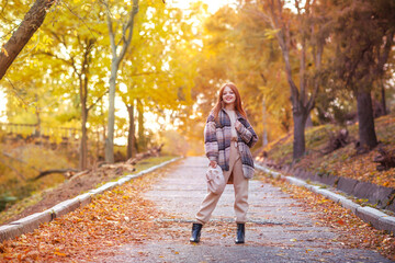 A young beautiful girl with long red hair stands right in the middle of the road going into the distance in an autumn park. Autumn outfit, fallen leaves, walk, good mood sunlight concept