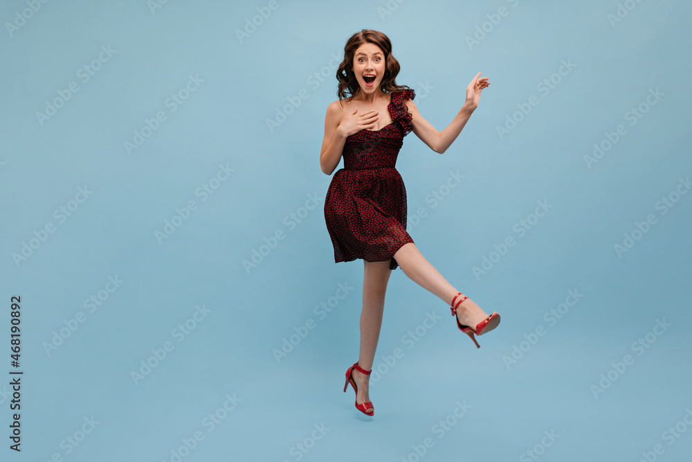 Poster Full-length gorgeous caucasian young lady bounces on blue studio background raising her leg. Happy brown-haired woman with wavy hair opened mouth, wearing short dress and red high-heeled shoes.