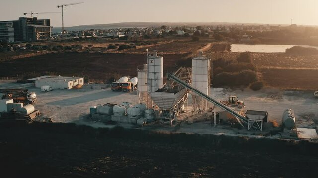 Stationary concrete plant. Cement mixer factory. Manufacturing, building, development. Storage reservoir silo towers in sunset light, rural background. Cinematic aerial drone flight zoom out panorama